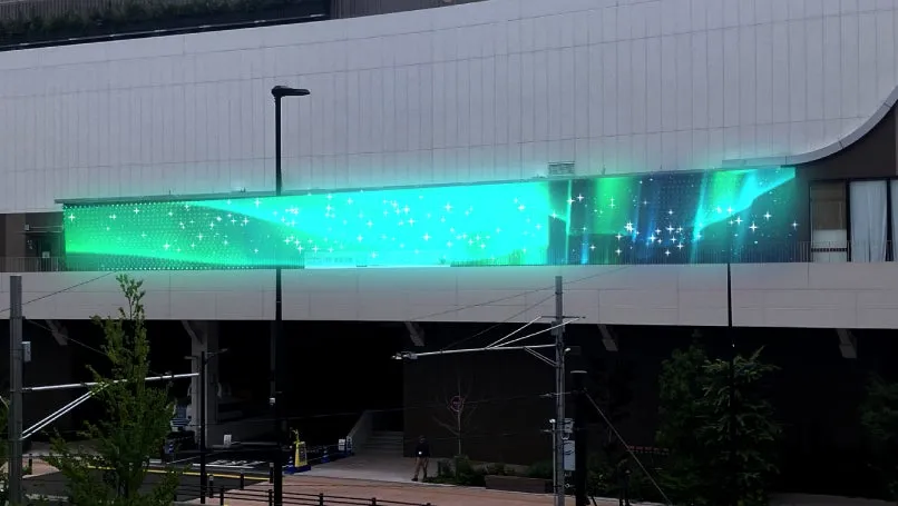 建物の壁に、オーロラのような光の演出がされている様子を捉えた写真です。緑と青のグラデーションが美しく、星のような光も散りばめられています。